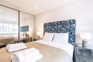 A hotel room with a large bed, a bedside table with a lamp, a mirror, and a window with a view of buildings. - Stylish Chelsea Flat Near Sloane Square (London)