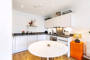 A modern kitchen with white cabinets, a round dining table, and a small orange side table. - Chic East London Stay With Outdoor Space (London)