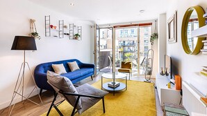 A modern living room with a blue sofa, a floor lamp, a coffee table, and a yellow rug. - Chic East London Stay With Outdoor Space (London)