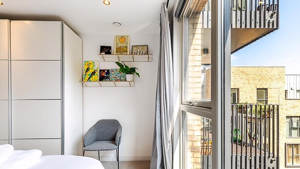 A bedroom with a bed, a chair, a window with a view of buildings, and shelves with pictures and a plant. - Chic East London Stay With Outdoor Space (London)