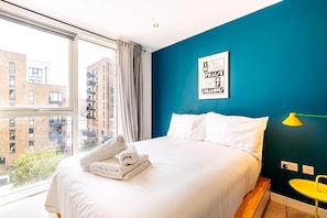 Ein Hotelzimmer mit großem Fenster, einem Bett mit weißer Bettwäsche, einer blauen Akzentwand und einem gerahmten Zitat an der Wand.