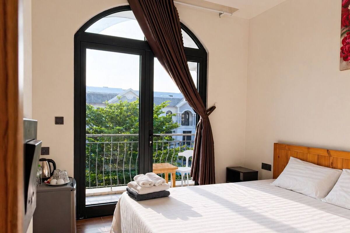Un dormitorio con cama, una mesita, una silla y un balcón con vistas a edificios y árboles.