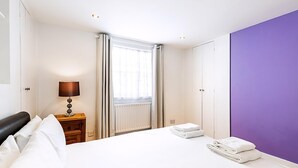 Une chambre d’hôtel avec un lit, une table de chevet avec une lampe, une fenêtre avec des rideaux et un mur d’accent de couleur violette.