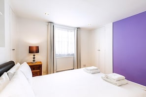 Ein Hotelzimmer mit Bett, Nachttisch mit Lampe, Fenster mit Vorhängen und einer lilafarbenen Akzentwand.