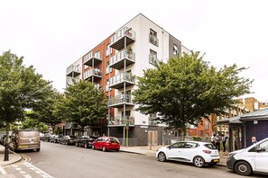 A multi-story residential building with balconies, parked cars, and trees along the street. - Modern Mile End Flat With Balcony Terrace (London)