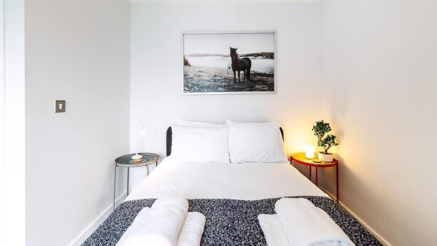 Ein Schlafzimmer mit einem Bett, Kissen, einem Nachttisch mit Lampe, einem gerahmten Bild an der Wand und einer kleinen Topfpflanze.