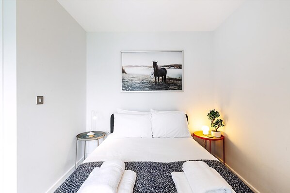 A bedroom with a bed, pillows, a bedside table with a lamp, a framed picture on the wall, and a small potted plant. - Modern Mile End Flat With Balcony Terrace (London)
