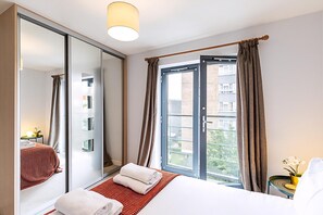 A hotel room with a bed, a large mirror, a window with curtains, and a view of buildings outside. - Modern Mile End Flat With Balcony Terrace (London)