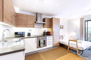 A modern kitchen with a built-in oven, a coffee maker, and a microwave. There is a small dining table and a bed with a wooden frame. - Modern Mile End Flat With Balcony Terrace (London)