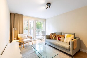 Ein modernes Wohnzimmer mit Sofa, Sessel, Glastisch und einem großen Fenster mit Vorhängen.