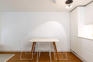 Ein schlicht eingerichteter Raum mit einem weißen Tisch und Stühlen, Holzboden und einer weißen Wand.