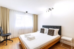 A hotel room with a bed, a chair, a bedside table, and a window with curtains. - Belgravia Living 2-bed, 1-bath Apartment With Balcony (London)