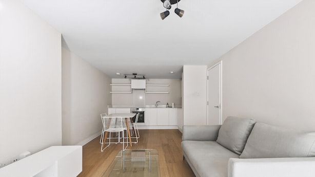 Uma sala de estar moderna com um sofá, uma mesa de centro de vidro e um espaço de jantar com uma mesa e cadeiras brancas. Uma cozinha com armários brancos e bancada é visível ao fundo.