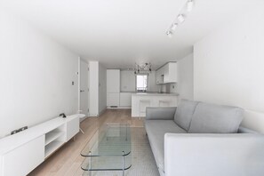 Ein modernes, minimalistisches Wohnzimmer mit einer Couch, einem Glastisch und einer Küchenzeile im Hintergrund.