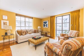 Uma sala de estar com sofá, poltrona, mesa de centro e uma janela grande com cortinas.