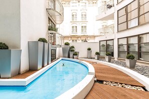 Ein Schwimmbecken mit Holzterrasse, umgeben von Blumentöpfen und einem weißes Gebäude mit Balkonen.
