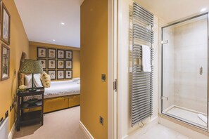 Quarto de hotel com uma cama, uma mesa de cabeceira com luminária, área de chuveiro com porta de vidro e parede com quadros.