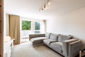 Ein modernes Wohnzimmer mit einem grauen Sofa, einem kleinen weißen Tisch und einem großen Fenster mit Vorhängen.