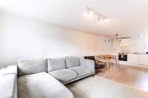 Ein moderner Wohnbereich mit einem grauen Ecksofa, einem Essbereich mit Holztisch und Stühlen sowie einer Küche mit weißen Schränken und einem Einbauofen.