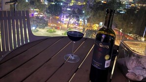 Une table avec une bouteille de vin, un verre et un bol de collations, sur un balcon donnant sur une vue panoramique de la ville la nuit.