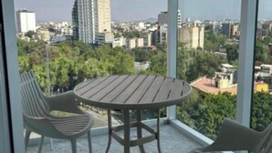 Un balcon avec une table ronde et deux chaises, donnant sur un paysage urbain composé d’immeubles et d’espaces verts.