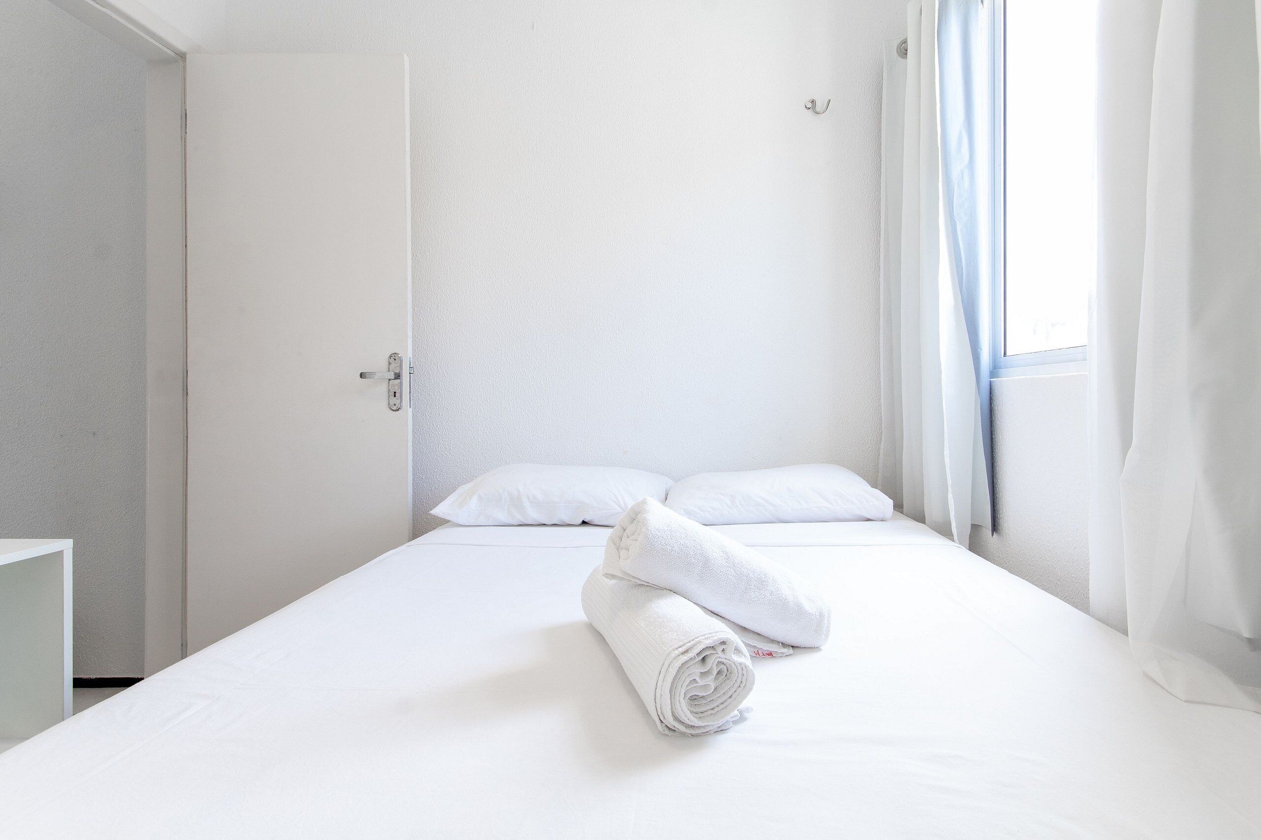 A hotel room with a bed, white linens, a rolled towel, a window with curtains, and a door.