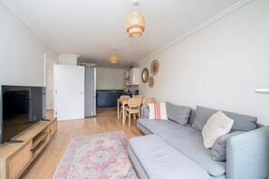 A living room with a grey sofa, a flat-screen TV on a wooden stand, a patterned rug, and a dining area with a table and chairs. - Bright 2-bed Apartment in North Acton (London)