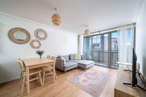 A modern living room with a dining table, grey sectional sofa, wicker wall decor, and a large window with a view. - Bright 2-bed Apartment in North Acton (London)