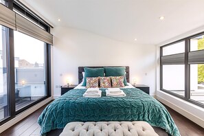 Um quarto com uma janela grande, uma cama com colcha verde-água, duas almofadas, um criado-mudo com abajur e um banquinho estofado.