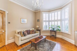 A living room with a sofa, a coffee table, a chandelier, a window with shutters, and a plant. - Charming Ground Floor Flat, West Kensington (London)