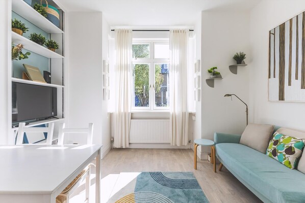 A modern living room with a sofa, a small table, a TV, and shelves with plants. - Stylish West London Stay by Hammersmith (London)