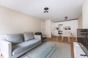 Ein modernes Wohnzimmer mit einem grauen Sofa, einem Glastisch und einer Küchenzeile mit weißen Schränken und einem Esstisch.