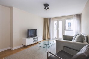 Ein Wohnzimmer mit Glastisch, einer Couch, einem Fernseher auf einem weißen Schrank und einem Fenster mit Vorhängen.