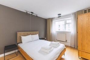 A hotel room with a bed, a nightstand, a wooden wardrobe, and a window with curtains. - 2BR Central Belgravia Retreat (London)