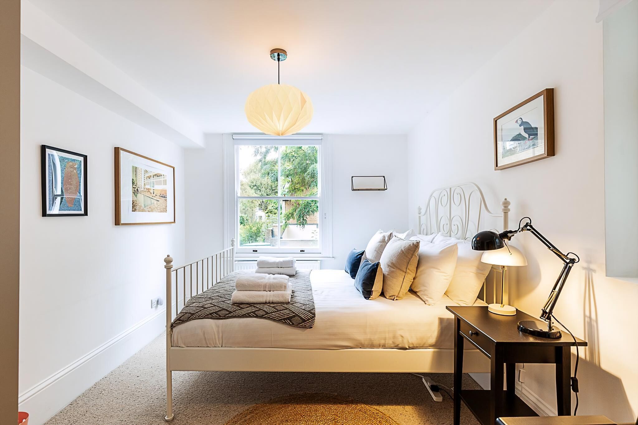 A bedroom with a bed, bedside table, lamp, window, and framed pictures on the wall.
