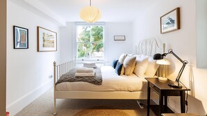 Un dormitorio con cama, mesita de noche, lámpara, ventana y cuadros en la pared.