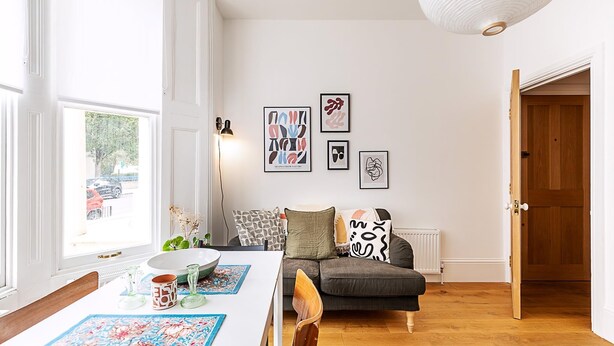 Una habitación con una mesa de comedor, un sofá y un radiador. Hay cuadros en la pared y una puerta a la derecha.