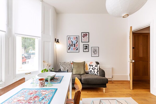 Ein Zimmer mit Esstisch, einer Couch und einem Heizkörper. An der Wand hängen Bilder in Rahmen und rechts ist eine Tür zu sehen.