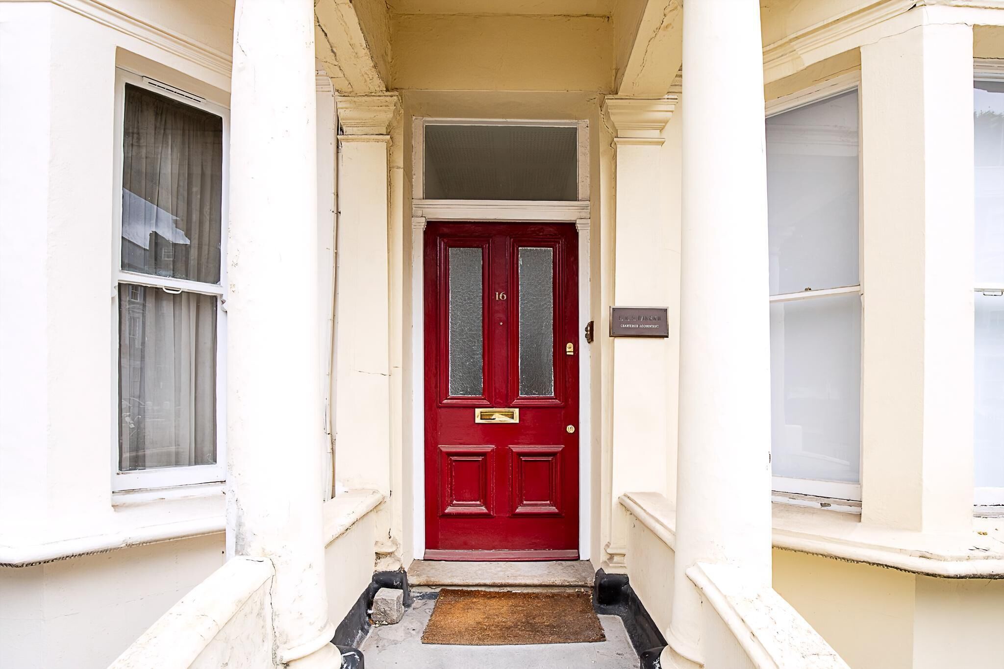 A red door with a number plate, a doormat, and a narrow entrance with white walls and columns.