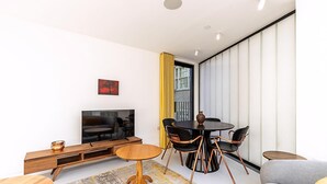 Un salon moderne comprenant un canapé, un téléviseur, une table à manger avec des chaises et une grande fenêtre avec des rideaux.