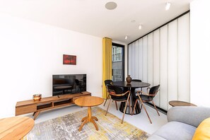 Ein modernes Wohnzimmer mit Sofa, Fernseher, Esstisch mit Stühlen und einem großen Fenster mit Vorhängen.