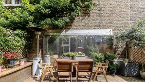 Eine Terrasse im Freien mit Backsteinmauer, Holztisch und -stühlen, Topfpflanzen und einer hängenden Leuchte.