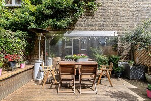 An outdoor patio with a brick wall, wooden table and chairs, potted plants, and a hanging light fixture. - Bright 3-bedroom Family Home Near Little Venice (London)