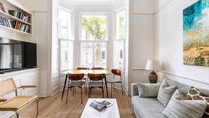 Ein Wohnzimmer mit Fernseher, Bücherregalen, einem Esstisch mit Stühlen, einer Couch und einem großen Fenster mit Rollläden.