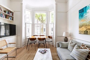 A living room with a television, bookshelves, a dining table with chairs, a sofa, and a large window with shutters. - Bright 3-bedroom Family Home Near Little Venice (London)