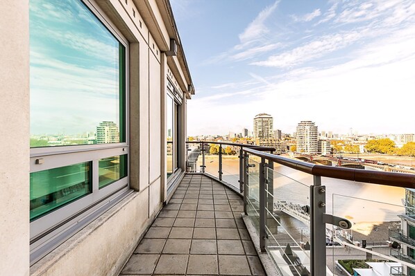 A balcony with a view of a river and cityscape, featuring a tiled floor and glass railings. - Riverside Luxury With Iconic Views (London)