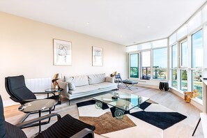 A modern living room with a sofa, chairs, a glass coffee table, and large windows offering a city view. - Riverside Luxury With Iconic Views (London)