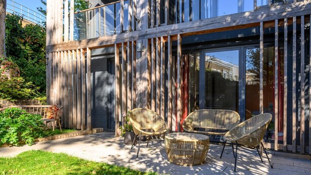 A modern building with large windows, a wooden fence, and outdoor seating with wicker chairs and a round table. - Stylish Family Retreat in Trendy Hackney (London)