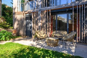 Un bâtiment moderne doté de grandes fenêtres, d’une clôture en bois et d’un espace extérieur aménagé avec des chaises en osier et une table ronde.