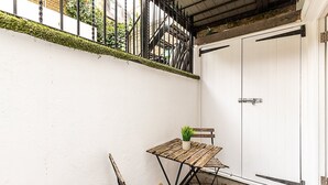 Un piccolo patio esterno con tavolo e panchetta in legno, una pianta in vaso e una porta bianca.